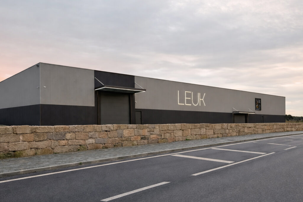 Fachada exterior del pabellón industrial de LEUK con muro de piedra y acceso por carretera.